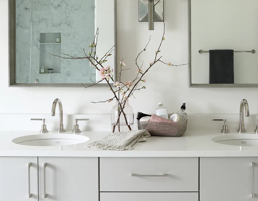 Modern white double vanity with chrome faucets, two mirrors, a floral vase, and countertop toiletries in a minimalist bathroom.
