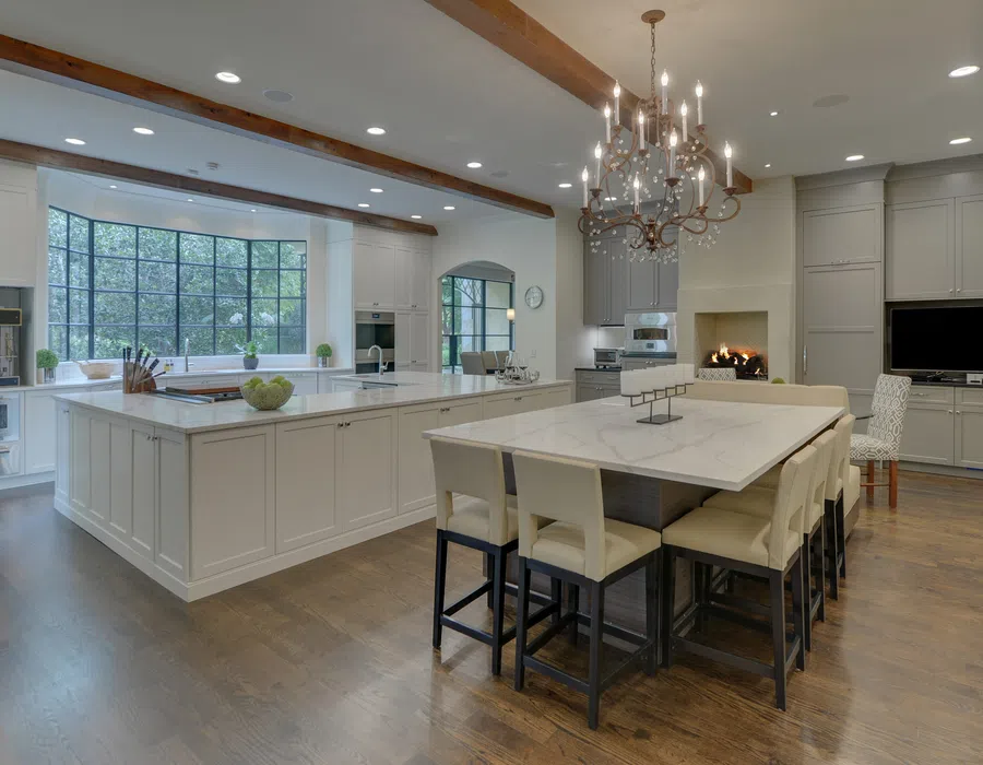 Modern spacious kitchen with large marble island, dining area, chandelier, wood beams, and wide windows overlooking greenery.